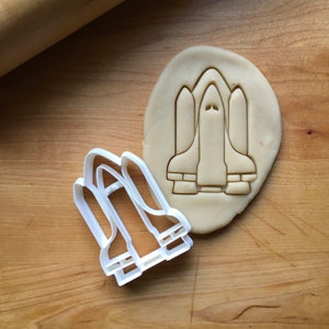 May include: A white plastic space shuttle cookie cutter next to a space shuttle-shaped cookie cut from dough. The cookie cutter is on a wooden surface.