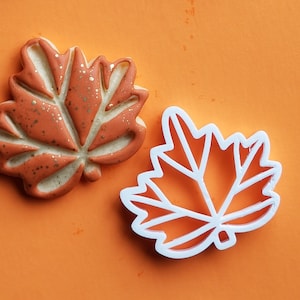 May include: Two leaf-shaped cookie cutters, one orange and one white. The orange cutter has a detailed design with a speckled texture. The white cutter has a simple, geometric design.