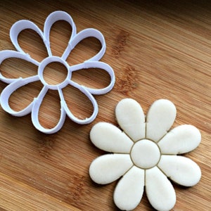 May include: A white daisy-shaped cookie cutter next to a daisy-shaped cookie on a wooden surface. The cookie cutter is made of plastic and has a simple design. The cookie is light-colored and has a similar shape to the cutter.