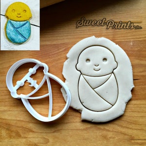 May include: A white plastic cookie cutter in the shape of a baby swaddled in a blanket. A sample cookie is shown next to the cutter, made from unbaked dough.