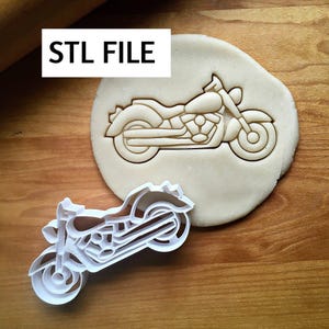 May include: A white plastic motorcycle-shaped cookie cutter beside a motorcycle imprint in dough. The text "STL FILE" is in a white rectangle in the upper left corner. The image is on a wooden surface.