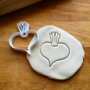 May include: A white plastic cookie cutter in the shape of a beet, next to a piece of dough with the beet shape pressed into it. The cutter has a heart-shaped body with a leafy top. The dough is on a wooden surface.