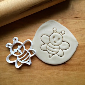 May include: White plastic cookie cutter in the shape of a cartoon bee. The cutter is shown next to a bee-shaped impression in a piece of white dough.