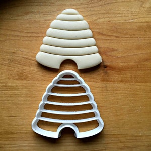 Pu&ograve; includere: Un tagliapasta in plastica bianca a forma di alveare con la parte superiore arrotondata e il fondo piatto. Il tagliapasta presenta una serie di creste per creare un motivo a nido d'ape. Un alveare di pasta da biscotti bianca &egrave; mostrato accanto al tagliapasta.