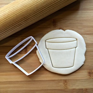 May include: A white plastic cookie cutter shaped like a plant pot next to a cut-out plant pot shape in dough. A wooden rolling pin is in the background. The items are on a wooden surface.