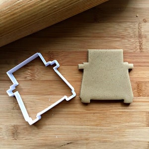 May include: A white plastic cookie cutter in the shape of a toaster sits next to a cut-out cookie on a wooden surface. A wooden rolling pin is in the background. The cookie is light brown.