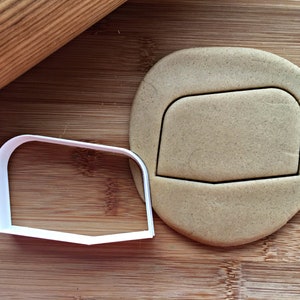 May include: A white metal cookie cutter in the shape of a rectangle with rounded corners and a beveled bottom edge, next to a cut-out cookie. A wooden rolling pin is in the background. The cookie and cutter are on a wooden surface.