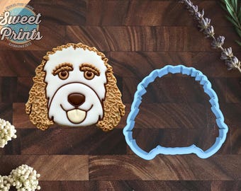 May include: A dog-shaped cookie decorated with white icing and tan textured fur details, featuring brown eyes, a dark nose, and a tongue, alongside a light blue plastic cookie cutter in the same shape, with the Sweet Prints logo visible.