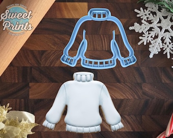 May include: A light blue plastic sweater-shaped cookie cutter positioned above a matching white sweater-shaped cookie with a high collar and ribbed cuffs. The text Sweet Prints Elevate your baking Established 2015 is visible.
