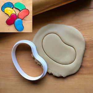 May include: A white plastic cookie cutter shaped like a jelly bean, with a piece of cookie dough pressed into it. The cookie cutter is on a wooden surface, next to a rolling pin.