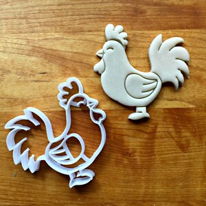 May include: A white plastic rooster-shaped cookie cutter and a matching beige dough cutout of a rooster. The cookie cutter features a detailed outline of the bird with a comb, wattle, and tail feathers, while the dough shows the same shape.