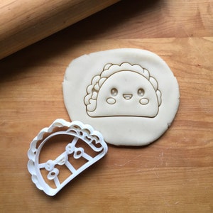 May include: White plastic cookie cutter shaped like a cartoon taco with a smiling face. The cutter is on a white surface with a cutout of the taco shape.