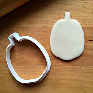 May include: A white plastic pumpkin cookie cutter and a plain white pumpkin-shaped cookie dough on a wooden surface.