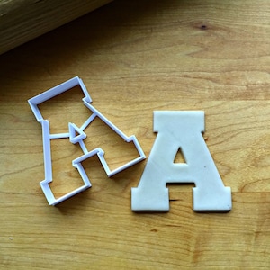 May include: Two white plastic cookie cutters in the shape of the letter A. One cutter is a solid shape and the other is an outline.