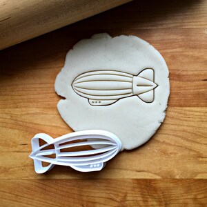 May include: A white plastic cookie cutter shaped like an airship or blimp. The cutter is on a wooden surface with a piece of dough that has been cut out with the cutter.