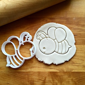 May include: White plastic cookie cutter shaped like a cartoon bee. The cutter is shown on a wooden surface with a rolled out piece of dough that has been cut with the cutter.
