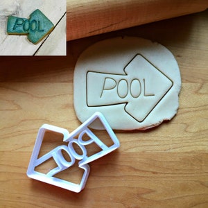 May include: A white plastic cookie cutter in the shape of an arrow with the word "POOL" inside. The cutter is next to a cutout of the same shape in dough, and a finished cookie with the word "POOL" is in the upper left corner.