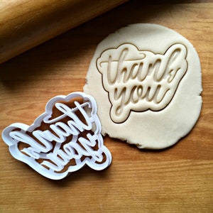 May include: Two white plastic cookie cutters on a wooden surface. One cutter is shaped like the words "Thank You" and the other is shaped like the words "Thank You Always".