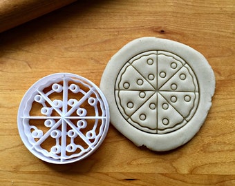 May include: A white plastic pizza-shaped cookie cutter next to a piece of dough with a pizza design pressed into it. The cutter has a circular shape with cut-out sections to form the pizza slices and toppings. The dough shows the pizza design.
