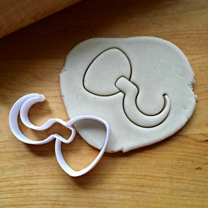 May include: A white plastic cookie cutter and a cut-out shape in a piece of dough. The cutter is in the shape of a heart with a curved handle. The dough is on a wooden surface.