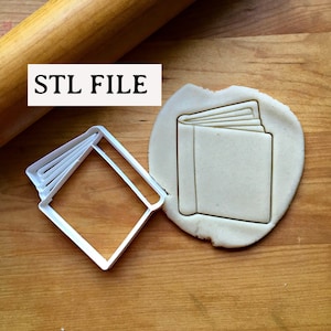 以下が含まれることがあります： 閉じた本の形をした白いプラスチック製のクッキーカッター。カッターは木製の表面にあり、カッターで切り抜かれた生地の隣にあります。画像には「STL FILE」というテキストが表示されています。