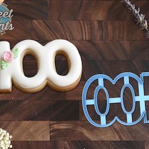 May include: A number 100 shaped cookie with smooth white icing and a small pink rose with green leaves, alongside a light blue plastic cookie cutter in the same number 100 shape. The logo Sweet Prints Established 2015 is in the top left corner.