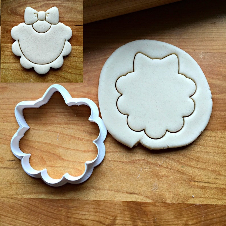 May include: A white plastic cookie cutter shaped like a bib with a bow on top. The cutter is on a wooden surface next to a piece of dough that has been cut out with the cutter.