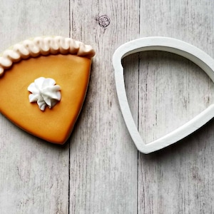 May include: A white plastic cookie cutter shaped like a pie slice, next to a decorated sugar cookie in the shape of a pie slice with a white frosting swirl on top.