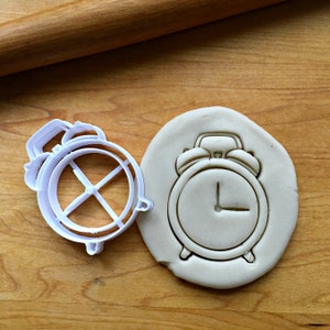 May include: A white plastic alarm clock cookie cutter next to a cookie with an alarm clock imprint. The cookie cutter is on a wooden surface with a rolling pin in the background. The cookie has the time set at 1:00.