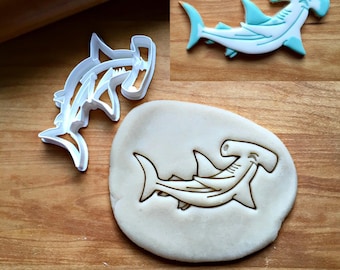 May include: A white plastic hammerhead shark cookie cutter, a piece of dough with a hammerhead shark impression, and a finished hammerhead shark cookie decorated with white and light blue icing, all featuring the distinct hammerhead shape.