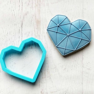 K&ouml;nnte beinhalten: Ein hellblauer herzf&ouml;rmiger Ausstecher neben einem dekorierten Keks. Der Keks ist hellblau mit einem geometrischen Design. Der Ausstecher ist aus Kunststoff. Der Keks hat einen hellbraunen Rand.