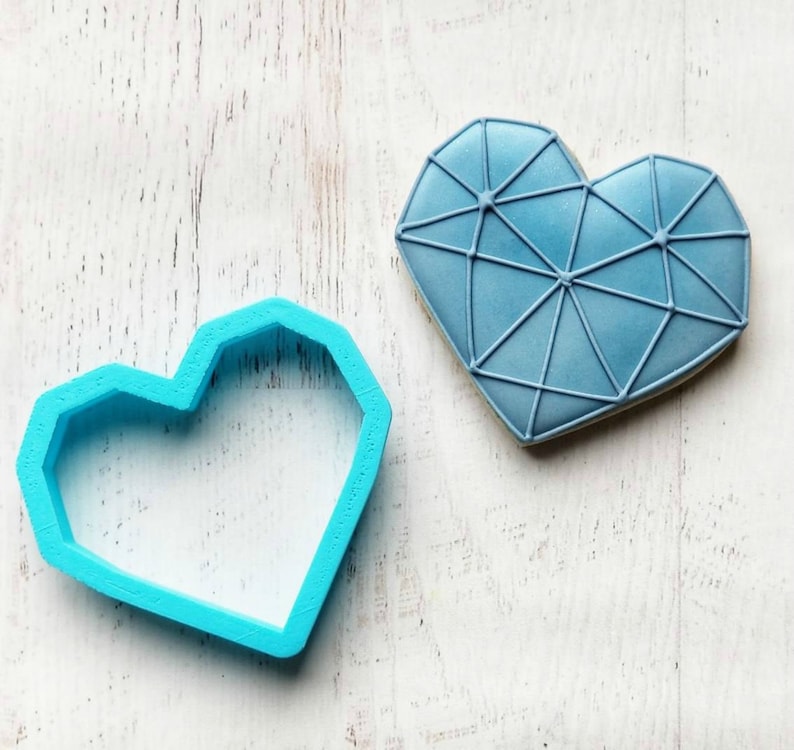 K&ouml;nnte beinhalten: Ein hellblauer herzf&ouml;rmiger Ausstecher neben einem dekorierten Keks. Der Keks ist hellblau mit einem geometrischen Design. Der Ausstecher ist aus Kunststoff. Der Keks hat einen hellbraunen Rand.