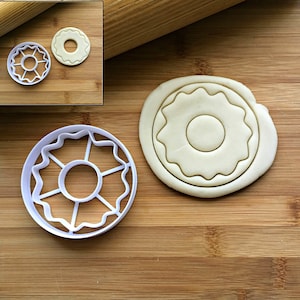 May include: White plastic cookie cutter shaped like a donut with a scalloped edge. A plain donut cookie is shown next to the cutter.