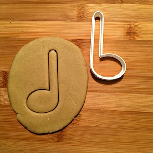 May include: A musical note cookie cutter and a cookie cut in the shape of a musical note sit on a wooden surface. The cookie cutter is white plastic, and the cookie is a light brown color.