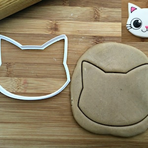 May include: A white plastic cat-shaped cookie cutter next to a piece of rolled dough with a cat-shaped impression. In the top right corner, a finished cookie is decorated with white icing, pink inner ears, black eyes, and black whiskers.