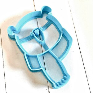 May include: A light blue plastic cookie cutter shaped like a bear wearing a scarf. The bear has a simple design with a round head, two ears, and a small nose.