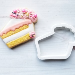 May include: A decorated sugar cookie shaped like a slice of cake, with pink frosting, sprinkles, and a small pink flower. Next to it is a white plastic cake-shaped cookie cutter.
