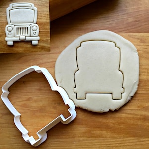 May include: A white plastic cookie cutter in the shape of a vintage truck, next to a cutout of the same shape in dough. The truck has a rectangular body, round headlights, and a detailed grill.