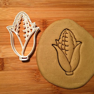 May include: White plastic cookie cutter shaped like an ear of corn. The cutter is shown on a wooden surface with a brown cookie dough imprint of the cutter.