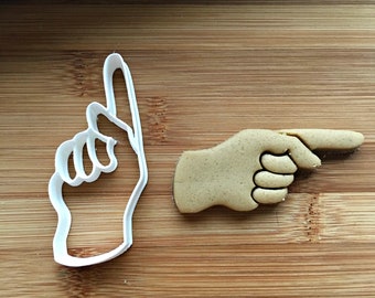 Può includere: Un tagliapasta di plastica bianca a forma di mano che punta verso l'alto, e un biscotto marrone a forma di mano che punta verso destra.