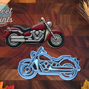 May include: A motorcycle-shaped cookie decorated with red and silver icing, featuring a black seat and detailed engine components, positioned above a light blue plastic motorcycle-shaped cookie cutter on a dark wood surface.