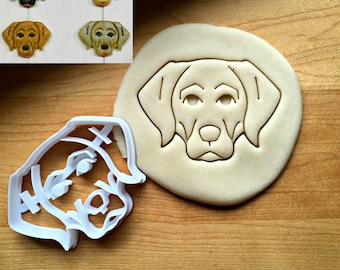 May include: A white plastic cookie cutter in the shape of a dog's head. The cookie cutter is on a wooden surface. There are also several cookies in the shape of a dog's head on the surface.