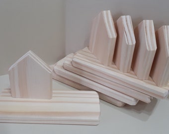 May include: A set of unfinished wooden house-shaped ornaments. The ornaments are stacked in a pile, with one ornament standing upright on a wooden base.