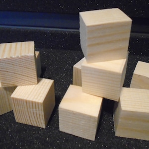 May include: A group of eight unfinished wooden cubes, stacked and scattered on a black surface. The cubes are made of natural wood and have a smooth, unpainted finish.