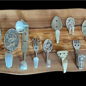 May include: A selection of handcrafted metal hooks and hangers, each uniquely shaped from repurposed silverware. The hooks are mounted on a wooden board with a natural grain, showcasing a rustic and vintage aesthetic. The hooks vary in size and design.