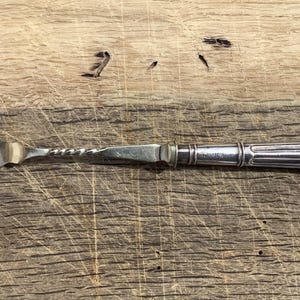 Pode incluir: Um garfo de cocktail em metal prateado com uma cabeça de três pontas, uma secção central torcida e um cabo cilíndrico com detalhes estriados verticais. O utensílio tem um acabamento polido e um design fino e alongado.