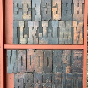 May include: A selection of vintage wooden letterpress printing blocks, displayed in a red tray. The blocks feature uppercase letters and numbers in a bold, serif font. The wood has a natural, aged appearance with variations in colour.
