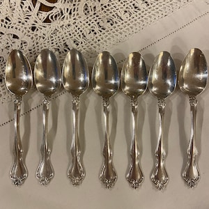 May include: Eight silver spoons with ornate handles. The spoons are arranged in a row on a white crocheted tablecloth.