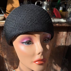 May include: A black, woven hat with a rounded crown and a delicate veil. The hat is displayed on a mannequin head with painted features, including pink eyeshadow and red lipstick. The hat's texture is visible, and it appears to be made of a straw-like material.