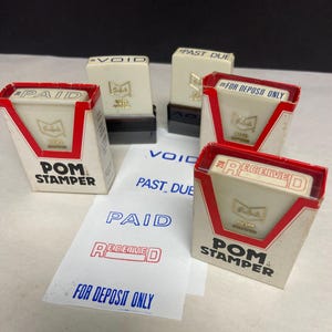 Puede incluir: Una colección de sellos vintage "POM Stamper" con embalaje rojo y blanco. Los sellos están etiquetados como "PAID", "RECEIVED", "VOID" y "FOR DEPOSIT ONLY". Los sellos están sobre una superficie blanca con papel impreso.